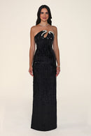 A woman modeling the Étincelle gown in black: a strapless paillette sequin column dress with an asymmetric neckline and a sculpted crystal accent across the bust, against a plain light beige studio background.