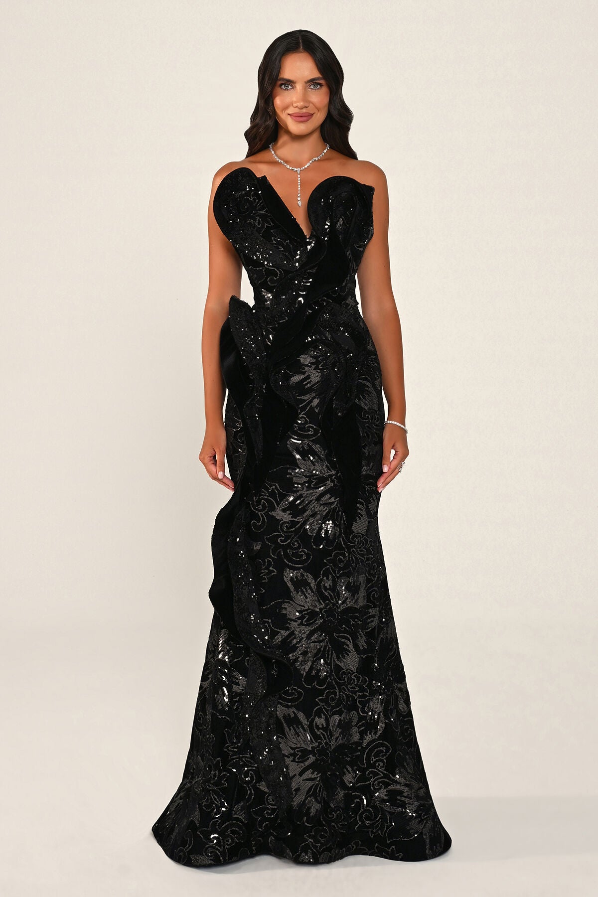A woman modeling the Ombrelle gown in black: a strapless velvet mermaid dress with a deep sweetheart neckline, a sculpted asymmetric velvet ruffle and a floral sequin skirt, against a plain light beige studio background.