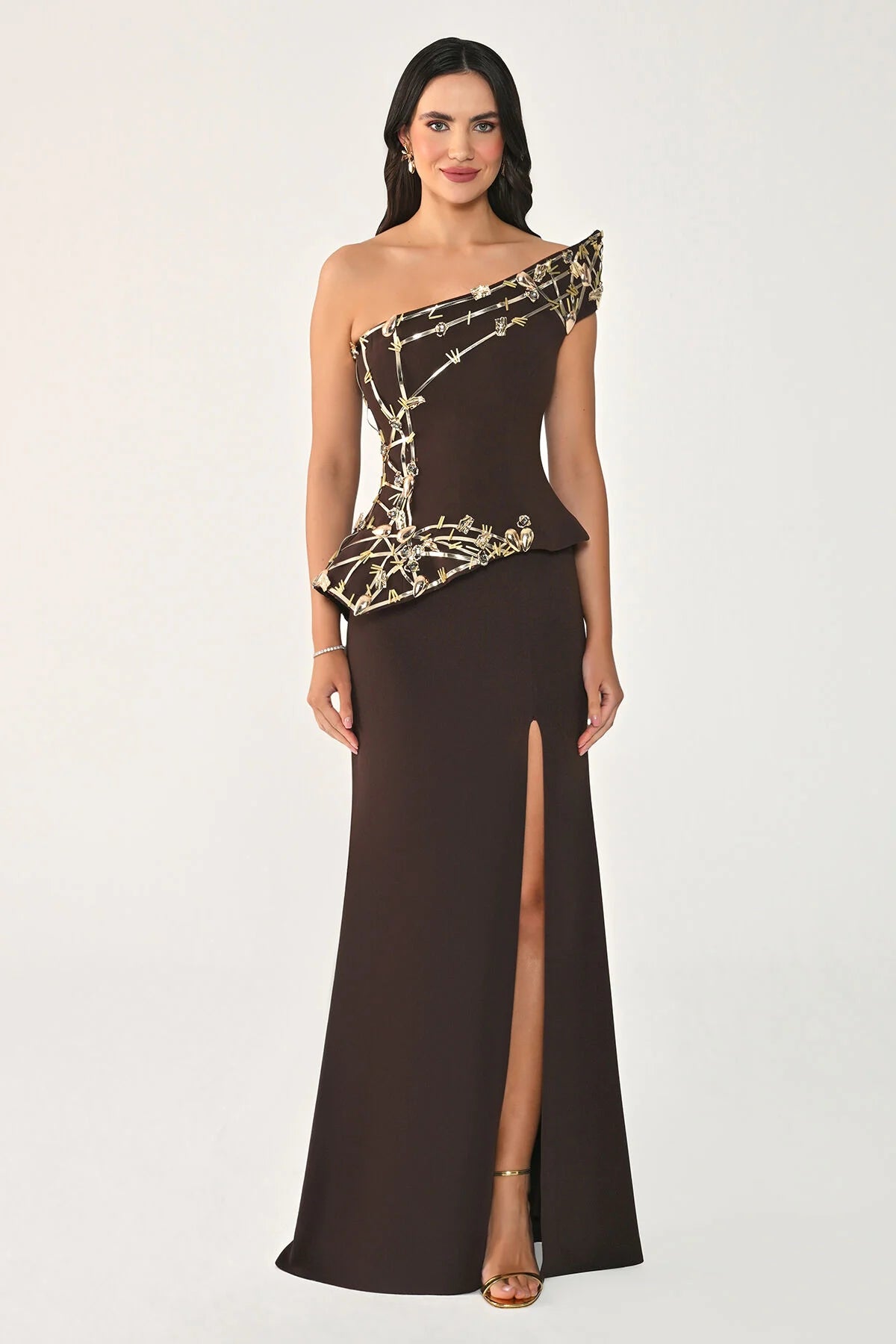 A woman modeling a floor-length one-shoulder evening gown in dark brown with metallic gold geometric embroidery on the bodice and upper skirt, photographed against a plain light gray studio background.