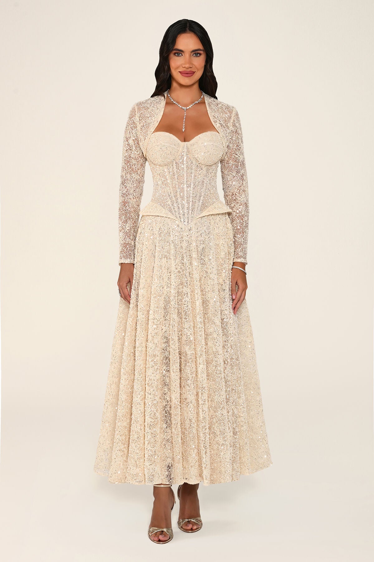 A woman modeling the Clarisse dress in beige: a sequin floral lace midi dress with a corset bodice, sweetheart neckline and matching long-sleeve bolero, against a plain light beige studio background.