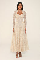 A woman modeling the Clarisse dress in beige: a sequin floral lace midi dress with a corset bodice, sweetheart neckline and matching long-sleeve bolero, against a plain light beige studio background.