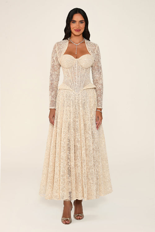A woman modeling the Clarisse dress in beige: a sequin floral lace midi dress with a corset bodice, sweetheart neckline and matching long-sleeve bolero, against a plain light beige studio background.